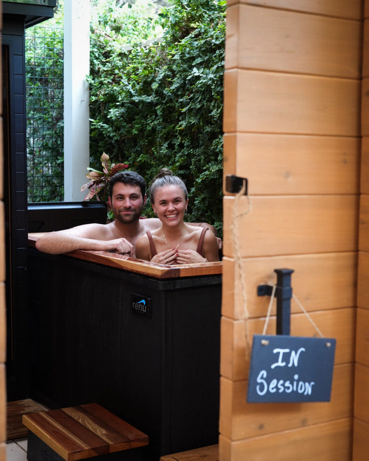 Two people in a cold plunge with a 'In Session' sign on a wooden wall.