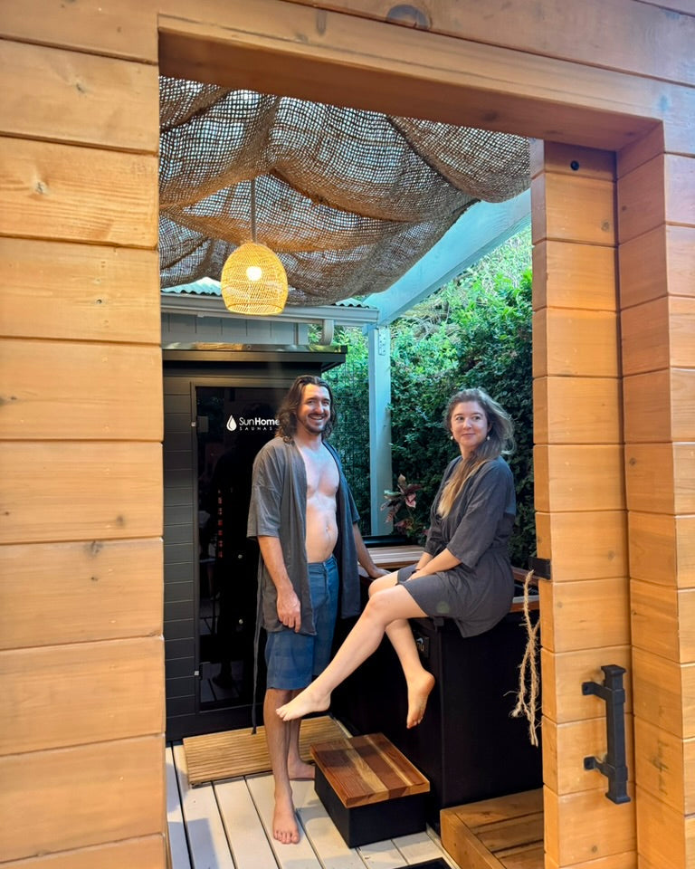 Two people standing in a wooden outdoor shower area with a modern sauna and cold plunge